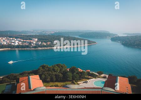 Vista aerea della costa frastagliata, dove le acque blu si incontrano con le lussureggianti isole verdi e l'antica città, una barca che attraversa la superficie scintillante Foto Stock