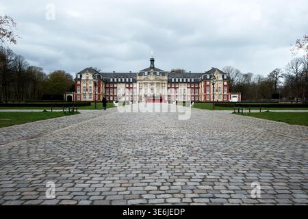 Muenster, Germania - 30 marzo 2025: Ampia vista del castello di Muenster con la piazza acciottolata e un autobus rosso di fronte all'edificio barocco sotto le nuvole Foto Stock