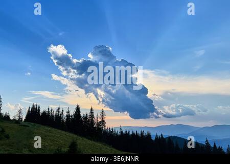 La grande e suggestiva formazione di nuvole nel cielo serale getta ombre sul tranquillo paesaggio montano. Alti pini si stagliano contro l'orizzonte e il cielo passa dal blu alle sfumature di arancione e giallo. Foto Stock