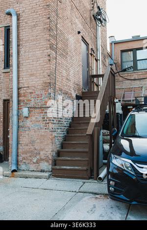 Una serie di scale di legno conduce ad una porta sul retro di un edificio in mattoni in una città. Un'auto nera è parcheggiata vicino alle scale. Tubi e linee di utilità Foto Stock