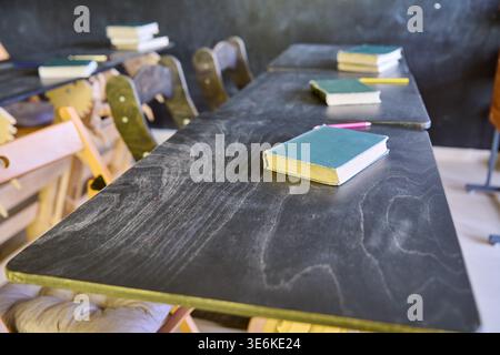 Scrivanie nere in legno con vecchi libri e matite consumati sparsi in aule vuote e con luce soffusa, con sedie rustiche e sfondo di lavagna Foto Stock