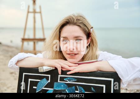 Pittrice donna adulta con capelli biondi, pelle chiara e lentiggini leggeri si appoggia su opere d'arte astratte sulla spiaggia. Lo sfondo calmo del mare e il cavalletto aggiungono un tocco creativo Foto Stock