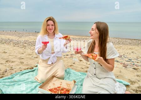 Due giovani donne condividono un rilassante picnic in riva al mare, sorridendo con bicchieri da vino rosa e fette di pizza sulla coperta. L'oceano calmo e il cielo coperto sono tranquilli Foto Stock