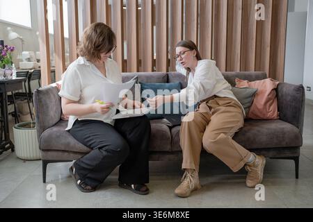 Due donne sedute vicino e che analizzano documenti sul divano insieme in un moderno ufficio aziendale. Analisi, documenti, donne, divano, moderno, affari. Foto Stock
