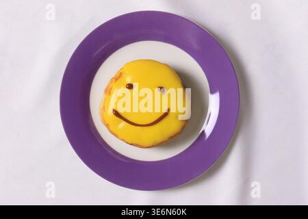 Morbida ciambella ripiena di crema ricoperta da glassa gialla brillante e un allegro design del viso sorridente, servita su un piatto viola e bianco su una tovaglia bianca. Foto Stock