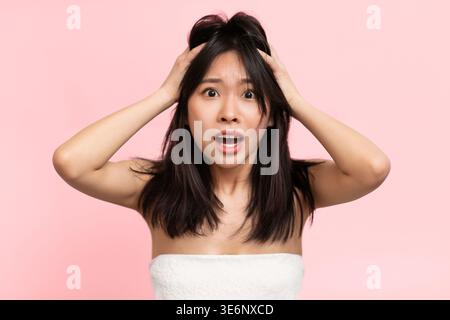 Donna con capelli scuri lunghi mostra sorpresa mentre tiene la testa davanti allo sfondo rosa Foto Stock