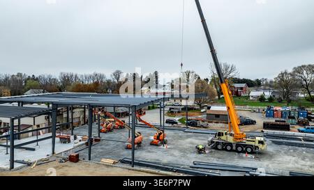 I lavoratori sono impegnati in un cantiere edile. Una grande gru solleva le travi in acciaio in posizione per un nuovo edificio. Sono presenti varie macchine e veicoli e alcune persone stanno guidando il lavoro. Foto Stock
