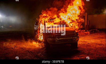 Un furgone va a fuoco in un'area buia di notte. Le fiamme eruttano dal veicolo man mano che il fumo riempie l'aria. Le luci vicine creano uno sfondo luminoso. Potrebbero essere presenti soccorritori di emergenza. Foto Stock