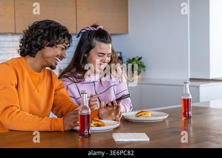 Coppie diverse che ridono e guardano lo smartphone all'isola della cucina, condividendo panini e bottiglie Foto Stock