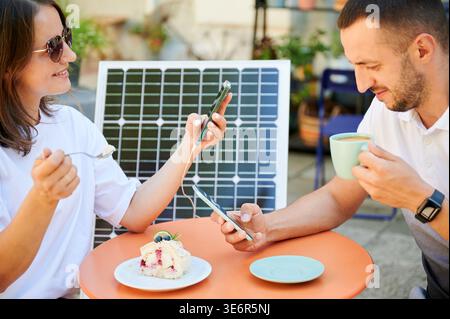 Uomo e donna che caricano lo smartphone dal pannello solare e si rilassano al bar. Coppia simpatica che utilizza un caricatore solare portatile per i dispositivi. Incantevole coppia seduto sul terrazzo sullo sfondo di una batteria solare. Foto Stock
