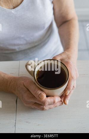 Donna anziana in camicia bianca che tiene in mano una tazza in ceramica calda con entrambe le mani rughe su un tavolo di legno bianco, routine tranquilla Foto Stock
