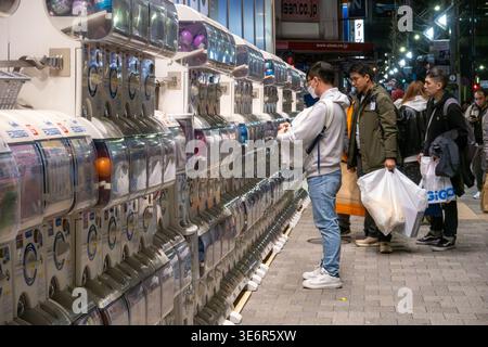 Una lunga fila di distributori automatici di capsule giocattolo, noti come gachapon, fa la fila di un marciapiede nel quartiere Akihabara di Tokyo, in Giappone, di notte. I pedoni curiosano e fanno shopping lungo la strada trafficata nel popolare quartiere dell'elettronica e degli anime. I distributori automatici di giocattoli a capsula, noti come gachapon, distribuiscono piccoli giocattoli da collezione in capsule sigillate con un elemento a sorpresa randomizzato. Presenti in tutto il Giappone nei portici, nei negozi e nelle stazioni ferroviarie, sono popolari sia tra i bambini che tra gli adulti per la loro novità e varietà. Foto Stock