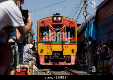 SAMUT SONGKHRAM, TAILANDIA-Mar 28, 2026: Treno diesel che passa dal mercato degli ombrelli pieghevoli alla piattaforma, atmosfera del mercato ferroviario, vista frontale Foto Stock