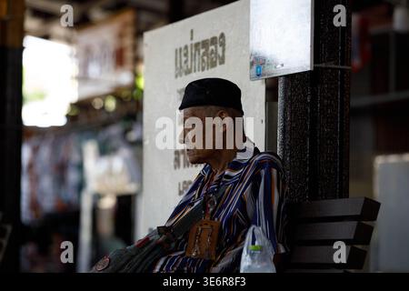 SAMUT SONGKHRAM, THAILAND-Mar 28, 2026: Anziano uomo che riposa sulla piattaforma ferroviaria di Maeklong, candida vita di strada tropicale, profilo cside Foto Stock