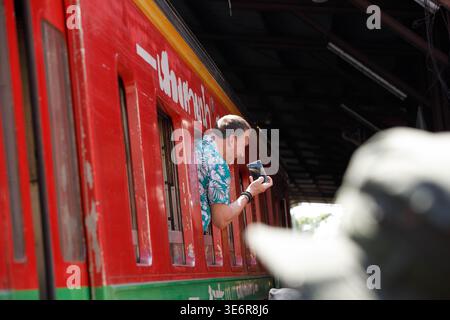 SAMUT SONGKHRAM, THAILAND-Mar 28, 2026: Turista maschio appoggiato fuori dalla carrozza ferroviaria per scattare foto, profilo laterale, al mercato di Maeklong. Foto Stock