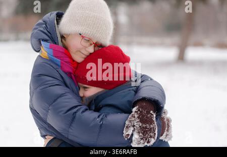Una ragazza che abbraccia calorosamente il fratello minore in un parco innevato, un tenero legame con i fratelli e il calore invernale della famiglia Foto Stock