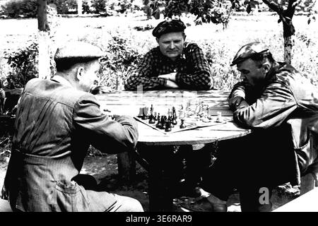Sloviansk, 1950 anni. Il territorio dell'impianto di armatura e isolamento (AIZ). Tre operai sovietici e membri del management si divertono con una partita a scacchi in un tavolo rotondo di legno sotto un albero prima di una dimostrazione pubblica. Questa autentica fotografia d'archivio in bianco e nero cattura il tempo libero all'aperto, il cameratismo e l'atmosfera rilassata tra i colleghi durante una festosa giornata primaverile. L'immagine storica d'epoca conserva splendidamente l'autentico stile di vita della classe operaia e il ricordo della tranquilla Donbas. Foto Stock