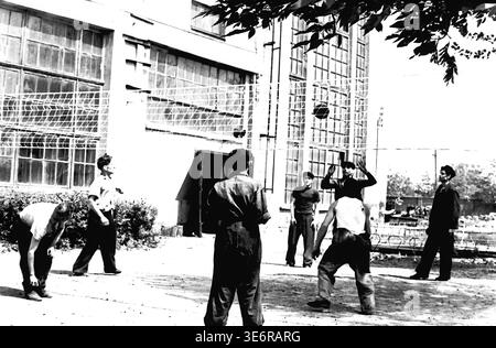 Sloviansk, 1950 anni. Il territorio dell'impianto di armatura e isolamento (AIZ). I lavoratori delle fabbriche sovietiche si divertono entusiasticamente con una vivace partita di pallavolo all'aperto nel cortile della fabbrica mentre si radunano per una prossima dimostrazione festiva. I giocatori sono posizionati in modo da ricevere la palla che passa sopra la rete sotto la luce del sole. Questa autentica fotografia d'archivio in bianco e nero cattura perfettamente l'importanza delle attività ricreative fisiche, del cameratismo sul posto di lavoro e dell'autentica atmosfera storica del tranquillo Donbas. Foto Stock