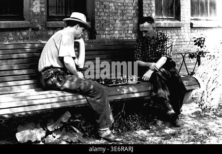 Sloviansk, 1950 anni. Il territorio dell'impianto di armatura e isolamento (AIZ). Due premurosi operai della fabbrica sovietica sono profondamente impegnati in un'intensa partita a scacchi mentre sono seduti su una lunga panchina di legno all'aperto prima di una festosa manifestazione pubblica. Un uomo che indossa un cappello elegante contempla con attenzione la sua prossima mossa strategica. Questa autentica fotografia d'archivio in bianco e nero cattura vividamente i popolari hobby intellettuali, il tempo libero all'aperto di tutti i giorni e la splendida e autentica storia della classe operaia nel tranquillo Donbas. Foto Stock