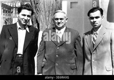 Sloviansk, 1950 anni. All'esterno dell'impianto di armatura e isolamento (AIZ). Tre fiduciosi uomini sovietici vestiti con abiti formali affilati posano insieme accanto a un tronco di albero grezzo nel cortile della fabbrica. I colleghi attendono con calma l'inizio di una manifestazione di massa festosa in una giornata molto luminosa e soleggiata. Questa fotografia d'archivio in bianco e nero altamente autentica mostra splendidamente la moda maschile della metà del secolo, le forti amicizie professionali, l'amministrazione della fabbrica e il ricco patrimonio culturale del pacifico Donbas. Foto Stock