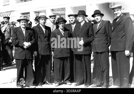 Sloviansk, 1950 anni. Il territorio dell'impianto di armatura e isolamento (AIZ). Un grande gruppo di sette dirigenti di fabbrica sovietici e ingegneri senior è orgoglioso di trovarsi in una lunga fila fuori da un edificio bianco. Gli uomini rispettabili indossano abiti formali, cappotti scuri e cappelli eleganti mentre si riuniscono per una manifestazione di massa festosa. Questa fotografia d'archivio espressiva in bianco e nero documenta perfettamente la leadership collettiva, la classica moda all'aperto della metà del secolo, gli eventi della comunità e la memoria storica duratura del pacifico Donbas. Foto Stock