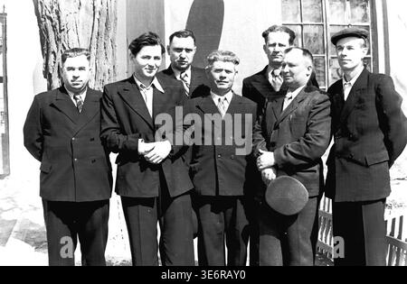 Sloviansk, 1950 anni. Il territorio dell'impianto di armatura e isolamento (AIZ). Sette importanti uomini sovietici, tra cui l'amministrazione della fabbrica e gli ingegneri anziani, posano con fiducia per un ritratto di gruppo vicino a un grande tronco di albero grezzo. Indossano abiti formali affilati e si godono il sole luminoso poco prima di partecipare a una manifestazione pubblica festosa. Questa fotografia d'archivio in bianco e nero altamente dettagliata documenta perfettamente i collettivi professionali del dopoguerra, la moda urbana maschile e le profonde radici culturali del pacifico Donbas. Foto Stock