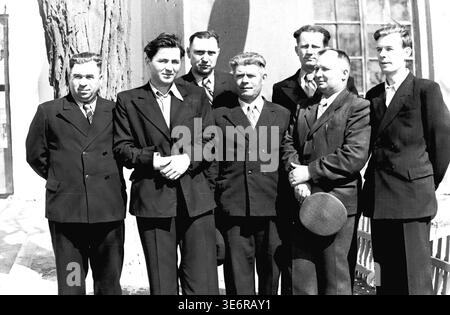 Sloviansk, 1950 anni. Il territorio dell'impianto di armatura e isolamento (AIZ). Sette importanti uomini sovietici, tra cui l'amministrazione della fabbrica e gli ingegneri anziani, posano con fiducia per un ritratto di gruppo vicino a un grande tronco di albero grezzo. Indossano abiti formali affilati e si godono il sole luminoso poco prima di partecipare a una manifestazione pubblica festosa. Questa fotografia d'archivio in bianco e nero altamente dettagliata documenta perfettamente i collettivi professionali del dopoguerra, la moda urbana maschile e le profonde radici culturali del pacifico Donbas. Foto Stock