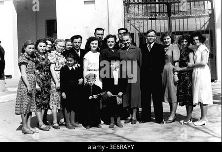 Sloviansk, 1950 anni. All'esterno dell'impianto di armatura e isolamento (AIZ). Un grande gruppo multigenerazionale di operai sovietici e le loro famiglie posa per un memorabile ritratto collettivo da un cancello di metallo decorativo prima di una festosa manifestazione di massa. I bambini sono felici di tenere piccole bandiere per le vacanze. Questa fotografia d'archivio in bianco e nero altamente autentica documenta brillantemente le riunioni della comunità della metà del secolo, la classica moda urbana, l'unità familiare e la storia culturale duratura del pacifico Donbas. Foto Stock