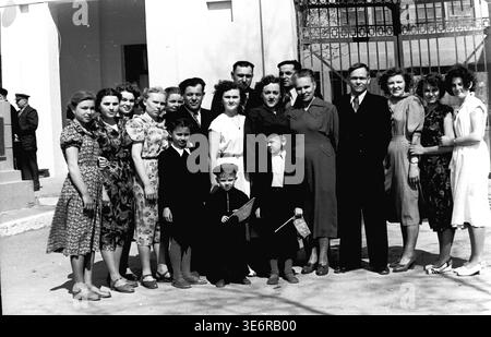 Sloviansk, 1950 anni. All'esterno dell'impianto di armatura e isolamento (AIZ). Un grande gruppo multigenerazionale di operai sovietici e le loro famiglie posa per un memorabile ritratto collettivo da un cancello di metallo decorativo prima di una festosa manifestazione di massa. I bambini sono felici di tenere piccole bandiere per le vacanze. Questa fotografia d'archivio in bianco e nero altamente autentica documenta brillantemente le riunioni della comunità della metà del secolo, la classica moda urbana, l'unità familiare e la storia culturale duratura del pacifico Donbas. Foto Stock
