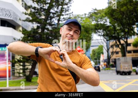 L'uomo con t-shirt arancione fa un gesto cardiaco con le mani sulla strada urbana. Ritratto dello stile di vita di un turista maschile che esprime amore e positività in città Foto Stock