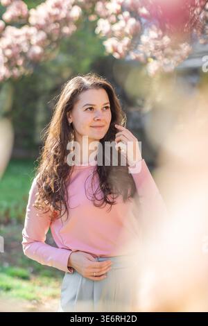 Giovane donna elegante in piedi sotto gli alberi di magnolia in fiore in un parco di Praga. Un'interpretazione moderna della moda, della natura e delle vibrazioni della città in primavera Foto Stock