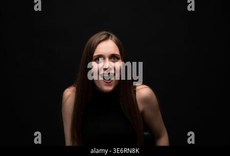 Giovane donna che mostra un'espressione di sorpresa ed eccitazione, con gli occhi larghi e la bocca aperta su uno sfondo scuro e isolato Foto Stock