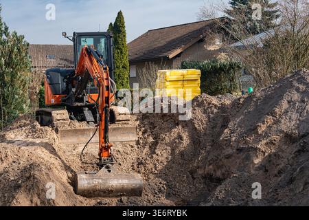 Un escavatore lavora in un cantiere. La macchina scava nel terreno vicino a una casa. I materiali da costruzione gialli sono impilati sul lato. Gli alberi lo sono Foto Stock