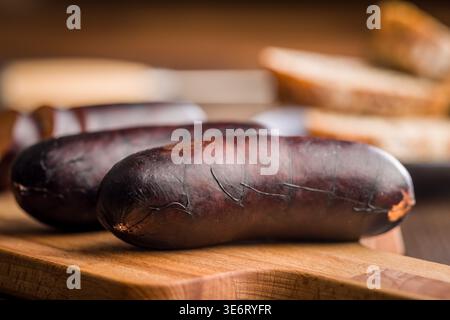La salsiccia di sangue affumicata, chiamata anche budino nero, si trova su un tagliere di legno con pane affettato offuscato dietro di esso, evocando rustico, saporito e mestiere Foto Stock