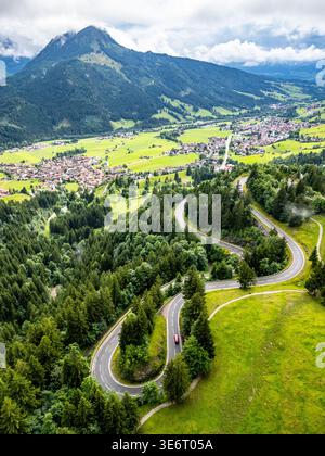 Una splendida vista aerea cattura le tortuose strade serpentine delle Alpi, circondate da lussureggianti colline verdi e vette montane. Ideale per gli amanti dell'avventura e dei viaggi su strada. Foto Stock