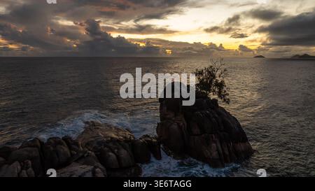 Roccia solitaria con palme tra le onde dell'oceano al tramonto. Seychelles, Mahe. Foto Stock