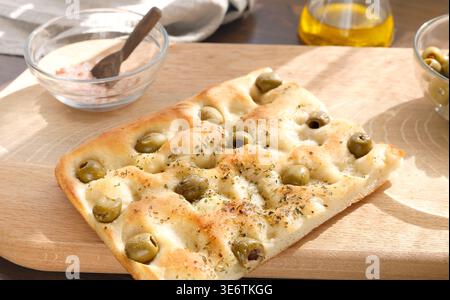 Tradizionale focaccia italiana con olive verdi ed erbe aromatiche su tagliere in legno. La dose ad angolo alto è dotata di sale in un recipiente di vetro e di una bottiglia di olio d'oliva Foto Stock