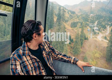 Il giovane ispanico con i capelli scuri siede in funivia, guardando le montagne e gli alberi durante una vacanza. Il paesaggio è caratterizzato da verdi colline e. Foto Stock