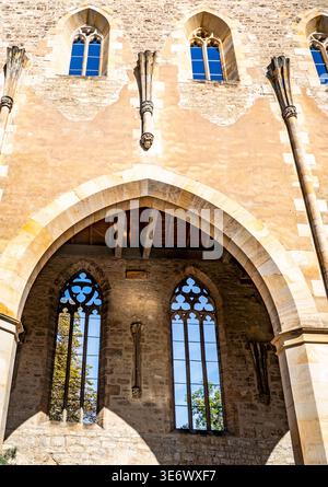 Facciata storica di un edificio in pietra con finestre gotiche ad arco e un grande arco arrotondato, con calde texture illuminate dal sole Foto Stock