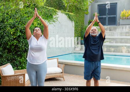 Coppie africane che fanno yoga sul patio piastrellato accanto alla piscina, indossano braccialetti e guardano Foto Stock