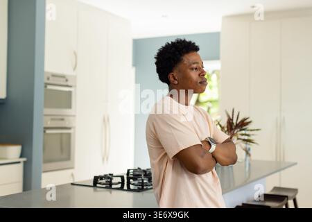 Uomo afro-americano in piedi con braccia incrociate da un'isola e fornello in smartwatch da cucina con tee rosa Foto Stock