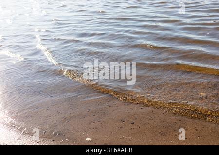 L'immagine mostra una superficie d'acqua tranquilla con onde dolci che si infrangono su una spiaggia sabbiosa, illuminata dalla luce del sole, creando un natu sereno e pacifico Foto Stock