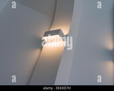 Dettagli architettonici precisi di una moderna applique quadrata che diffonde una calda luce soffusa contro una parete interna curva bianca liscia. La scena rappresenta e. Foto Stock