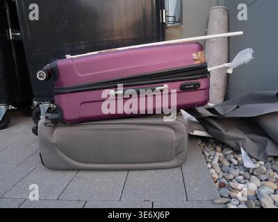 La valigia rigida viola poggia su una valigia grigia dai lati morbidi su una superficie pavimentata, con ciottoli e pareti grigie visibili sullo sfondo Foto Stock