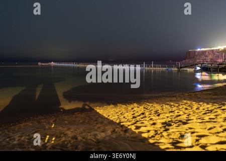 Vista notturna di un lungo molo in legno sulla spiaggia del Mar Rosso a Sharm El Sheikh, Egitto, con costa illuminata. Foto Stock