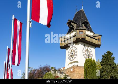 Vista dell'iconica torre dell'orologio contro un cielo azzurro limpido con bandiere austriache che sventolano dolcemente nella brezza, Graz, Stiria, Austria. Foto Stock