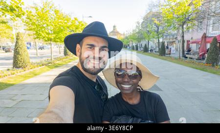 Coppia multiculturale felice che indossa cappelli e porta un gioioso Selfie mentre esplora una Scenic City Street Foto Stock