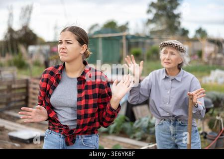 Due donne arrabbiate casual vicini di età diverse che discutono durante la stagione di orto il giorno di sole di primavera Foto Stock