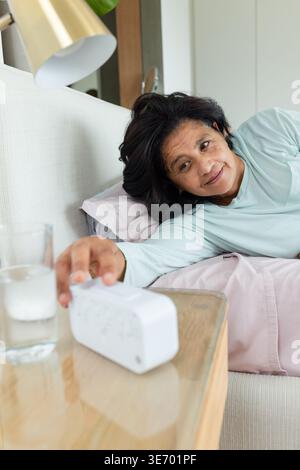 Donna africana matura sdraiata in camera da letto con la parte superiore azzurra che raggiunge la sveglia bianca Foto Stock