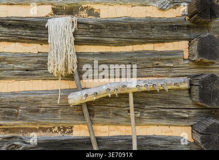 Vecchio fatto a mano attrezzi agricoli di rastrello in legno e la stringa mop appoggiata contro log cabin pareti Foto Stock